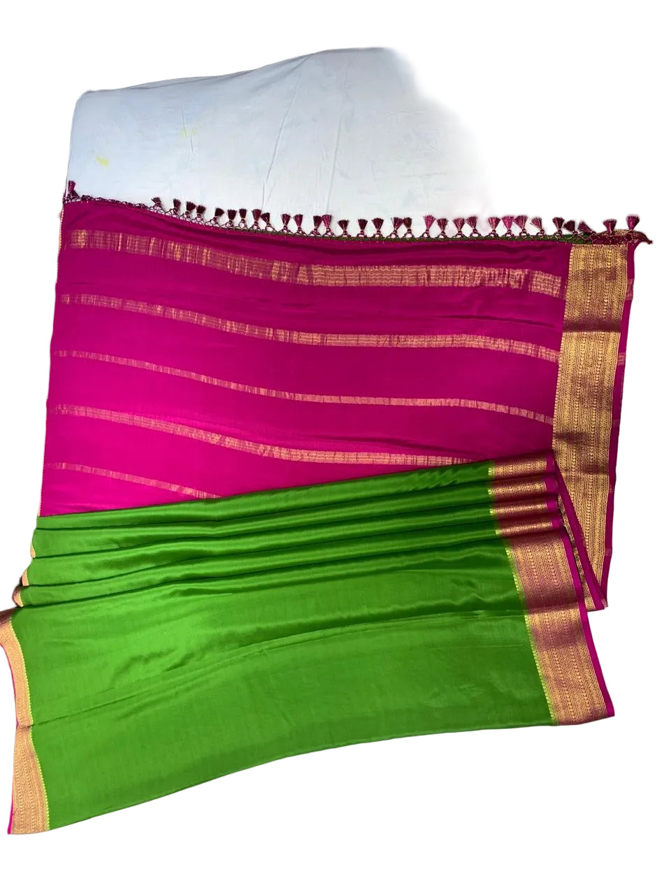 A folded and draped saree laid flat on a white sheet, featuring a bright magenta pink body with gold zari stripes, a green pallu section with gold border, and a pink tassel border.